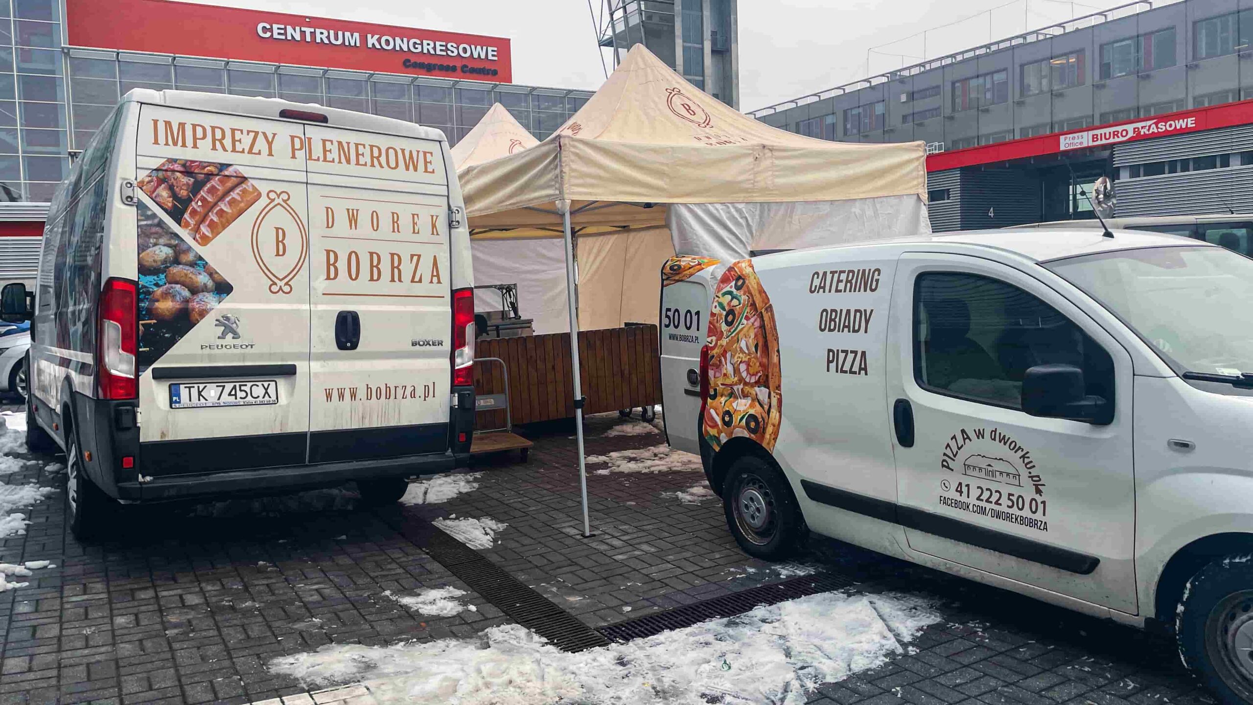 Stanowisko cateringowe na targach – namiot z grillem, firmowe samochody i obsługa dostarczająca jedzenie na halę.