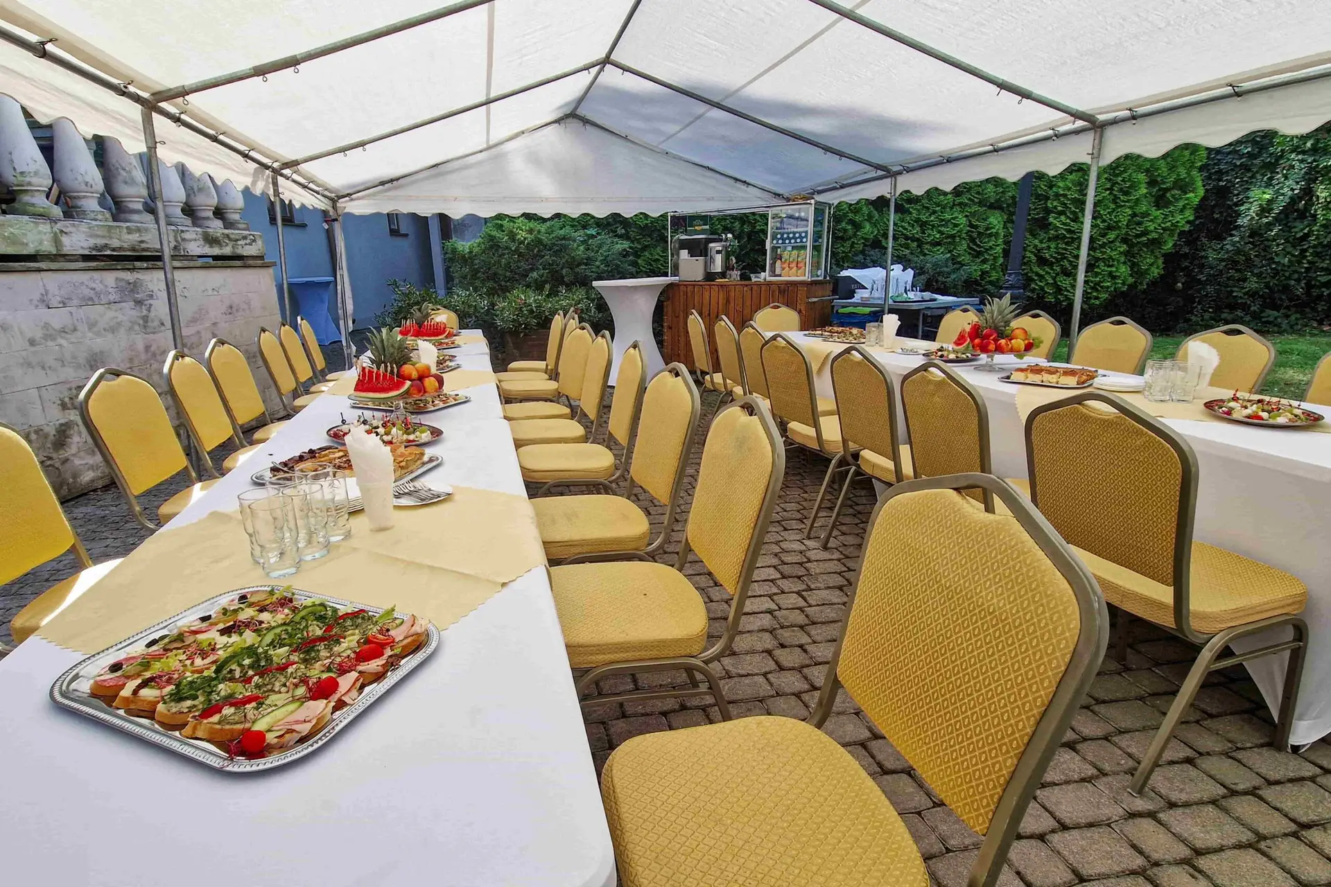 Plenerowy bankiet pod białym namiotem – stoły z przekąskami, owocami i kawowy barek z napojami.