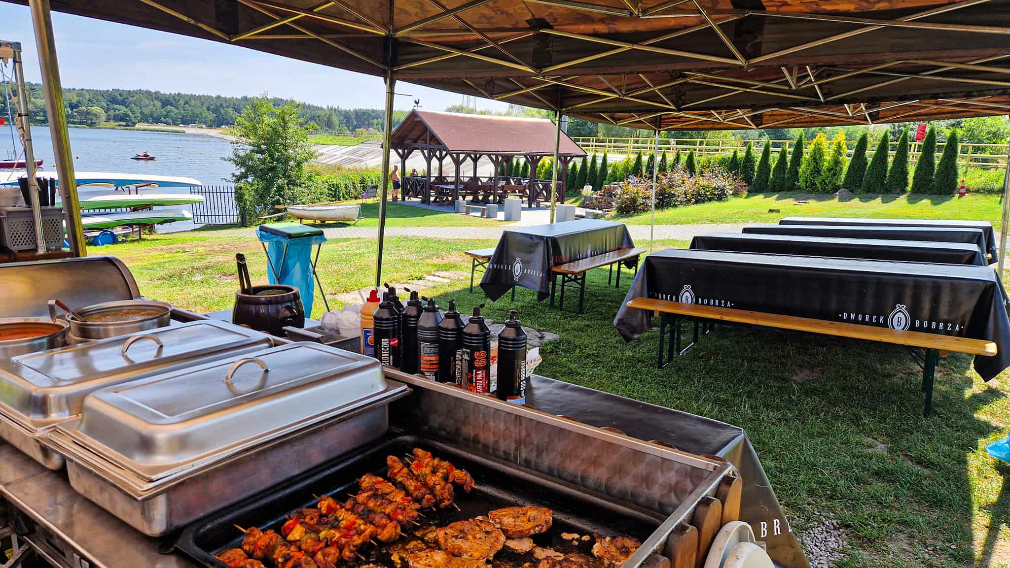 Grill na imprezie plenerowej – namioty, stoły i ławki, jedzenie i sosy, malownicza sceneria nad zalewem Cedzyna